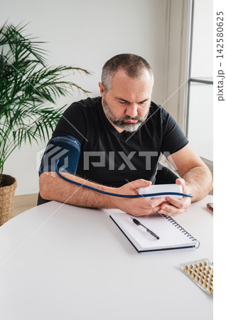 Worried Man Checking Blood Pressure with Home Monitor Vertical. Vertical photo of a worried mature man with an arm cuff looking at his digital home blood pressure monitor at a table, illustrating hype Worried Man Checking Blood Pressure with Home Monitor Vertical. Vertical photo of a worried mature man with an arm cuff looking at his digital home blood pressure monitor at a table, illustrating hype 142580625