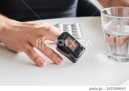 Close-up of Fingertip Pulse Oximeter Oxygen Reading. Close-up of a fingertip pulse oximeter showing the oxygen reading on a mature man's finger beside a glass of water and pills, illustrating home hea Close-up of Fingertip Pulse Oximeter Oxygen Reading. Close-up of a fingertip pulse oximeter showing the oxygen reading on a mature man's finger beside a glass of water and pills, illustrating home hea 142580643