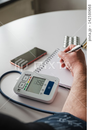 Close-up of Blood Pressure Monitor Reading Vertical. Vertical close-up of a digital home blood pressure monitor with a hand writing the reading in a notebook beside a smartphone, illustrating hyperten Close-up of Blood Pressure Monitor Reading Vertical. Vertical close-up of a digital home blood pressure monitor with a hand writing the reading in a notebook beside a smartphone, illustrating hyperten 142580668