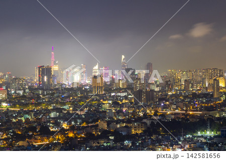 A panoramic urban skyline glows with colorful building lights beneath a dark, cloud-filled night sky. A panoramic urban skyline glows with colorful building lights beneath a dark, cloud-filled night sky. 142581656
