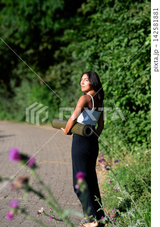 Woman in Woman Carrying a Yoga Mat in a Summer Park. A young Hispanic woman carries a rolled yoga mat on a path beside the calm lake on her way to an outdoor yoga session, embracing an active and mind Woman in Woman Carrying a Yoga Mat in a Summer Park. A young Hispanic woman carries a rolled yoga mat on a path beside the calm lake on her way to an outdoor yoga session, embracing an active and mind 142581881