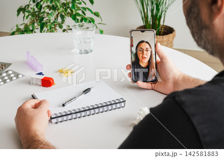 Man Speaking with Doctor on a Video Consultation. Mature man speaks with a female doctor on a smartphone video call at a table with a notebook and pill organizer, illustrating telemedicine, online con 142581883