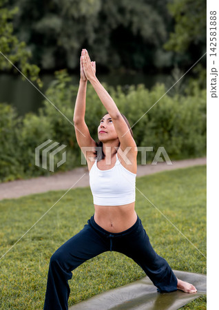 Fit Woman in Crescent Lunge With Prayer Hands on a Yoga Mat Outdoors. A young Hispanic woman practises a crescent lunge with prayer hands on the grass beside the water in the evening light, connecting 142581888