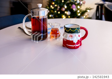 Santa Mug and French Press Tea with Honey by Christmas Tree. A red Santa Claus mug, a glass French press of tea and a small glass of honey on a white table before a glowing Christmas tree. Warm bokeh Santa Mug and French Press Tea with Honey by Christmas Tree. A red Santa Claus mug, a glass French press of tea and a small glass of honey on a white table before a glowing Christmas tree. Warm bokeh 142581943