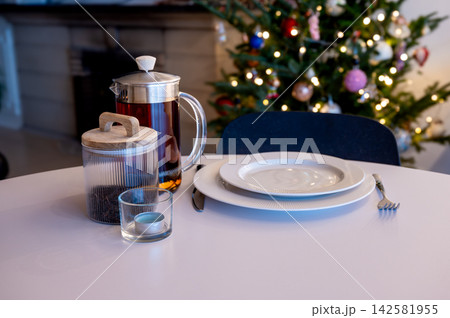 French Press of Tea with Festive Place Setting by Tree. A glass French press of tea beside white plates and a fork on a white table before a glowing Christmas tree with a fireplace. A cozy holiday tab French Press of Tea with Festive Place Setting by Tree. A glass French press of tea beside white plates and a fork on a white table before a glowing Christmas tree with a fireplace. A cozy holiday tab 142581955