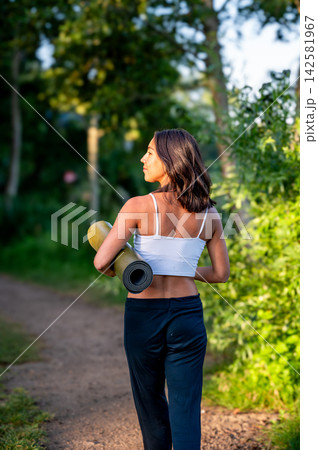Hispanic Woman in Woman Carrying a Yoga Mat in a Summer Park. A young Hispanic woman carries a rolled yoga mat in a lush green park in the evening light on her way to an outdoor yoga session Hispanic Woman in Woman Carrying a Yoga Mat in a Summer Park. A young Hispanic woman carries a rolled yoga mat in a lush green park in the evening light on her way to an outdoor yoga session 142581967
