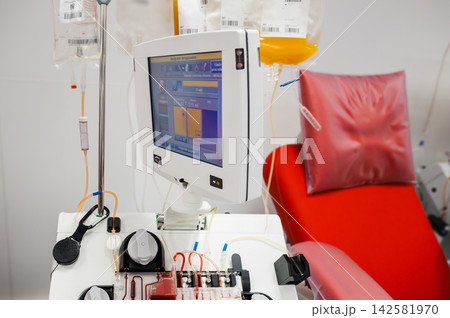 Apheresis Machine and Plasma Bags by Donation Chair, An apheresis machine with a screen and plasma bags on an IV stand beside a red donation chair at a blood bank, illustrating automated plasma collec Apheresis Machine and Plasma Bags by Donation Chair, An apheresis machine with a screen and plasma bags on an IV stand beside a red donation chair at a blood bank, illustrating automated plasma collec 142581970