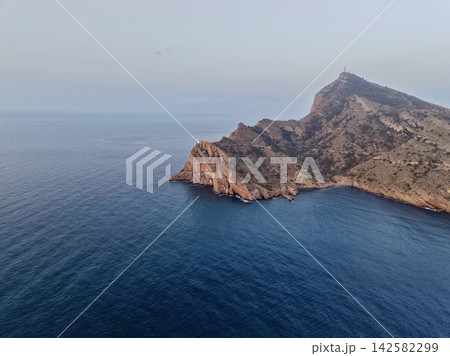 Rocky coastal headland with small lighthouse and communication mast above calm blue sea under pale sky, showing rugged cliffs, remote shoreline, and tranquil open horizon Rocky coastal headland with small lighthouse and communication mast above calm blue sea under pale sky, showing rugged cliffs, remote shoreline, and tranquil open horizon 142582299