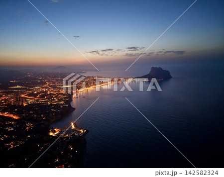 Twilight coastal city skyline with illuminated waterfront, calm reflective sea, clustered high-rise buildings, curved shoreline, and sea stack silhouette under evening clouds Twilight coastal city skyline with illuminated waterfront, calm reflective sea, clustered high-rise buildings, curved shoreline, and sea stack silhouette under evening clouds 142582324