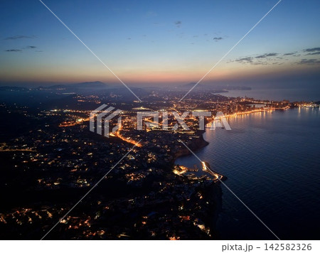 Coastal city lights tracing curved shoreline at twilight, with waterfront towers, calm reflective sea, distant hills, and soft evening sky over urban bay, with coast, city, light, shoreline detail 142582326