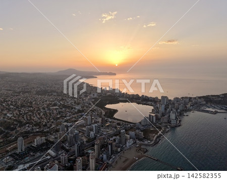 Coastal city skyline, bay, shoreline at sunrise with warm sun rays, calm sea, lagoon reflections, distant hills, glowing morning haze over harbor, with sunrise, coast, city, skyline detail 142582355