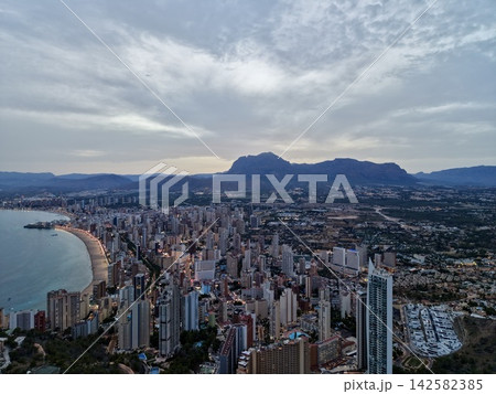 Coastal city skyline at dusk with mountain backdrop, illuminated waterfront, curved sandy bay, urban district, and dramatic cloudscape over calm seascape, with city, coastal, skyline, dusk detail 142582385