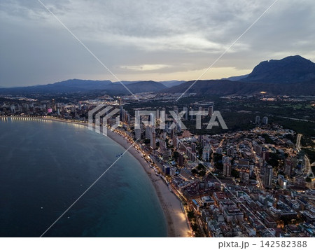 High angle cityscape at dusk showing illuminated beachfront highrises, curved sandy shore, waterfront promenade, calm sea and distant mountains beneath cloudy sky High angle cityscape at dusk showing illuminated beachfront highrises, curved sandy shore, waterfront promenade, calm sea and distant mountains beneath cloudy sky 142582388