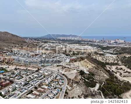 Coastal city skyline with residential hillside foreground, distant waterfront towers, dry terrain, winding road, and broad Mediterranean sea under pale blue sky 142582413