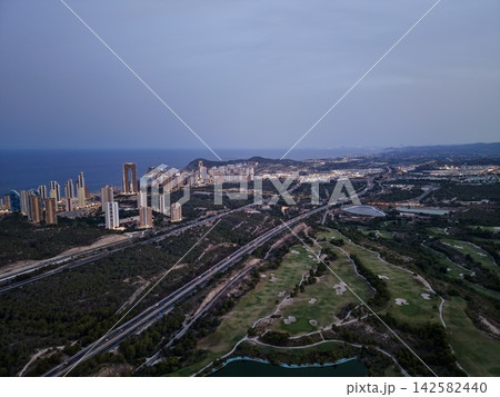 Coastal city skyline at dusk with illuminated highrise towers, shoreline views, winding traffic routes, forested hills, golf course, modern residential districts, and sea horizon Coastal city skyline at dusk with illuminated highrise towers, shoreline views, winding traffic routes, forested hills, golf course, modern residential districts, and sea horizon 142582440
