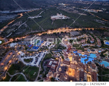 Twilight aerial view of coastal resort complex with glowing pools, lit terraces, tennis courts, pine hills, roads, parking areas, and seaside townscape under evening sky 142582447