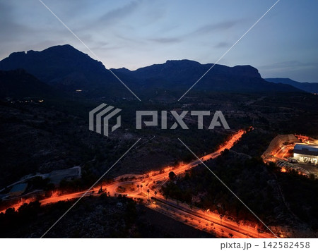 Mountain silhouette at dusk with glowing winding road, roundabout, streetlights, rugged ridge, sparse trees, and dramatic blue sky over quiet valley, with mountain, silhouette, dusk, twilight detail Mountain silhouette at dusk with glowing winding road, roundabout, streetlights, rugged ridge, sparse trees, and dramatic blue sky over quiet valley, with mountain, silhouette, dusk, twilight detail 142582458