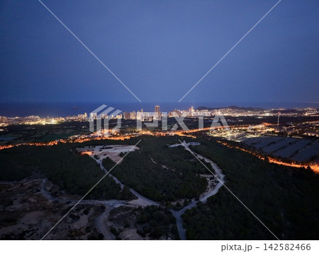 Coastal city skyline at dusk with illuminated towers, shoreline traffic, calm blue sea, dark hills, forested foreground, and glowing urban lights across waterfront Coastal city skyline at dusk with illuminated towers, shoreline traffic, calm blue sea, dark hills, forested foreground, and glowing urban lights across waterfront 142582466