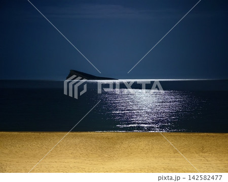 Moonlit seascape with distant rocky islet, calm reflective water, dark blue sky, soft shoreline sand, and quiet horizon creating minimalist nighttime coastal mood 142582477