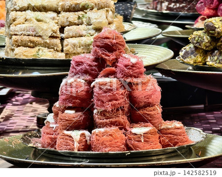 traditional middle eastern desserts stacked and displayed on trays in a culinary market stall with kunefe and baklava, red and beige colors, delicious and inviting mood, close-up from above. traditional middle eastern desserts stacked and displayed on trays in a culinary market stall with kunefe and baklava, red and beige colors, delicious and inviting mood, close-up from above. 142582519