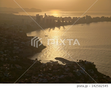 serene sunset reflection over coastal cityscape with golden and blue hues, aerial view of urban landscape featuring concrete, glass, and metal buildings and cliffs. serene sunset reflection over coastal cityscape with golden and blue hues, aerial view of urban landscape featuring concrete, glass, and metal buildings and cliffs. 142582545