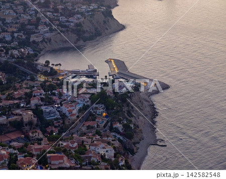 peaceful coastal town at dusk with houses, pools, and trees by the sea, aerial view from above, serene residential area by the sea, real estate and travel context, blue and white colors. 142582548