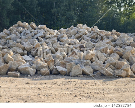 Large outdoor pile of gray rocks and boulders beside dirt ground with forest backdrop and sunlight, with rock, boulder, stone, gravel detail, showing pile, outdoor, yard, construction context Large outdoor pile of gray rocks and boulders beside dirt ground with forest backdrop and sunlight, with rock, boulder, stone, gravel detail, showing pile, outdoor, yard, construction context 142582784