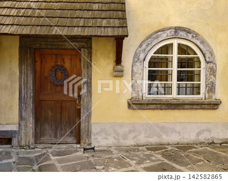 Weathered wooden door with wreath beside arched window on yellow stucco house, rustic courtyard with stone paving and historic charm, with door, wooden, doorway, wreath detail Weathered wooden door with wreath beside arched window on yellow stucco house, rustic courtyard with stone paving and historic charm, with door, wooden, doorway, wreath detail 142582865