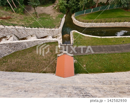 Aerial view over stone canal walls winding through parkland featuring green lawn, brick walkway, metal railings, curved water channel, and verdant trees on both sides, dramatic perspective Aerial view over stone canal walls winding through parkland featuring green lawn, brick walkway, metal railings, curved water channel, and verdant trees on both sides, dramatic perspective 142582930