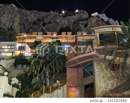 Night view hillside rocks illuminated terrace buildings along winding yellow railing stairway under dark sky overlooking coastal hillside resort, with night, hillside, rock, cliff detail Night view hillside rocks illuminated terrace buildings along winding yellow railing stairway under dark sky overlooking coastal hillside resort, with night, hillside, rock, cliff detail 142582988