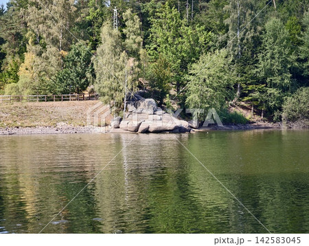 Sunlit lakeside shoreline featuring large rocks and boulders, dense pine forest, calm reflective water, green ripples, distant wooden railing, and tranquil natural scenery by shore in bright daylight Sunlit lakeside shoreline featuring large rocks and boulders, dense pine forest, calm reflective water, green ripples, distant wooden railing, and tranquil natural scenery by shore in bright daylight 142583045