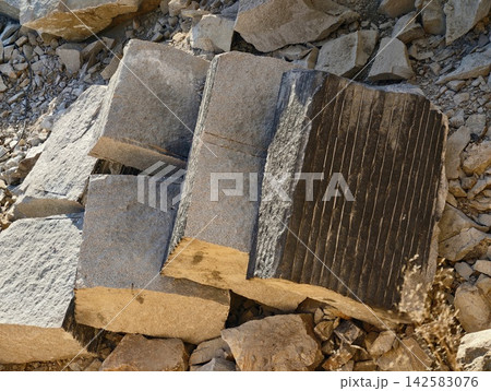Massive gray quarried stone blocks stacked on rugged rocky ground revealing angular faces, textured surfaces, industrial masonry scene, heavy construction material showcase Massive gray quarried stone blocks stacked on rugged rocky ground revealing angular faces, textured surfaces, industrial masonry scene, heavy construction material showcase 142583076