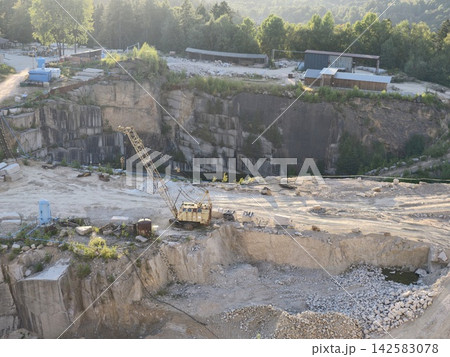 Quarry excavation site with steep rocky cliff, industrial machinery, and scattered rubble overlooking forested hillside at late day light, with quarry, excavation, cliff, rock detail 142583078