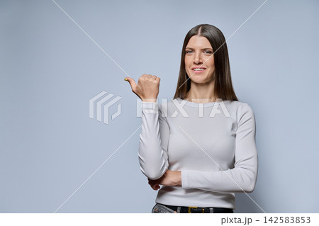 Portrait of young woman pointing thumb to copy space on grey background Portrait of young woman pointing thumb to copy space on grey background 142583853