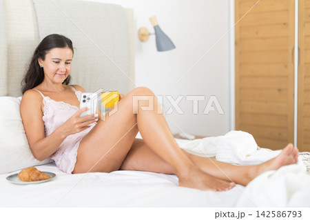 Woman drinking morning coffee and looking at smartphone screen in bedroom Woman drinking morning coffee and looking at smartphone screen in bedroom 142586793