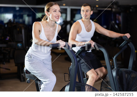 During classes on air bike, girl and man chat sweetly and smile During classes on air bike, girl and man chat sweetly and smile 142587255