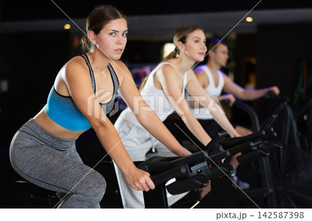Sportive young girl doing spin bike exercises in well-equipped gym Sportive young girl doing spin bike exercises in well-equipped gym 142587398