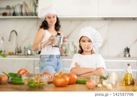 Little girl in chefs hat and white T-shirt stands in kitchen near table Little girl in chefs hat and white T-shirt stands in kitchen near table 142587706