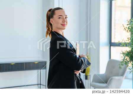 Confident business woman holding closed laptop in modern office Confident business woman holding closed laptop in modern office 142594420