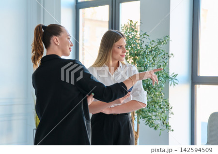 Two businesswomen standing during meeting pointing away in modern bright office Two businesswomen standing during meeting pointing away in modern bright office 142594509
