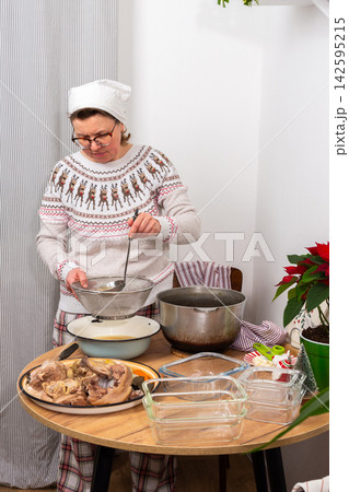 A woman stands in her kitchen preparing kholodets, aspic, a traditional Ukrainian dish. She strains the broth from the bones into a bowl while wearing comfortable clothes 142595215