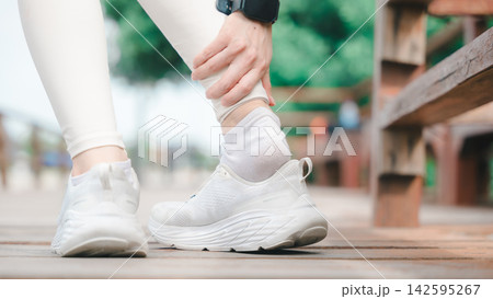 Female runner suffering achilles tendon injury holds painful ankle while training outdoors, active athlete experiencing sudden muscle pain or sprain during workout in public park 142595267