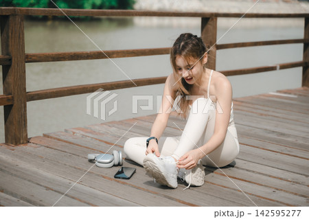 Female athlete ready for jogging on road as runner preparing for training in sport lifestyle, sitting on wooden boardwalk while tying shoelaces near smartphone and headphones 142595277
