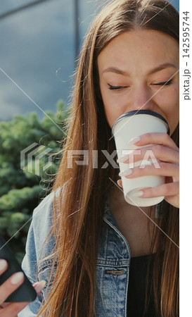 Vertical video. smiling woman typing message near office building during coffee break. Girl using smartphone and drinking coffee on the street bench. 142595744