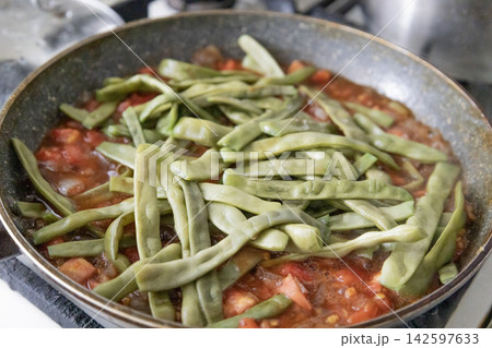 Fresh green beans cooking in a rich tomato sauce inside a pan. Close up view of homemade healthy vegetable stew preparation, organic food ingredients simmering for a delicious meal recipe. Fresh green beans cooking in a rich tomato sauce inside a pan. Close up view of homemade healthy vegetable stew preparation, organic food ingredients simmering for a delicious meal recipe. 142597633
