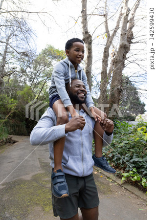 African American dad carrying son on shoulders walking paved park path in hoodies sneakers 142599410
