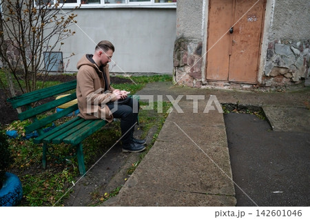 Young man sitting on a green bench absorbed in his smartphone in an urban environment on a chilly afternoon, surrounded by muted colors of tainted concrete Young man sitting on a green bench absorbed in his smartphone in an urban environment on a chilly afternoon, surrounded by muted colors of tainted concrete 142601046