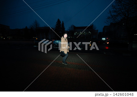 Man walks through a dark urban environment on a chilly evening, illuminated by streetlights and the glow of passing cars as the day gives way to night 142601048