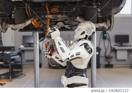 A humanoid robot with a multimeter examines the electrical wiring of a car chassis suspended on a lift in a workshop 142601117
