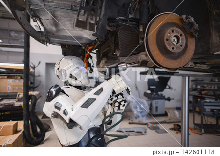 A humanoid robot with glowing blue eyes uses a power tool to repair the underside of a vehicle in a workshop setting A humanoid robot with glowing blue eyes uses a power tool to repair the underside of a vehicle in a workshop setting 142601118